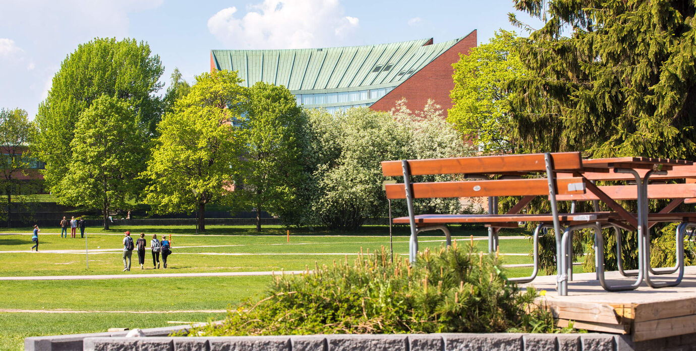 Puisto, jossa vihreitä puita ja nurmikkoa, penkkejä ja käveleviä ihmisiä. Taustalla rakennus, jossa vihreä katto.
