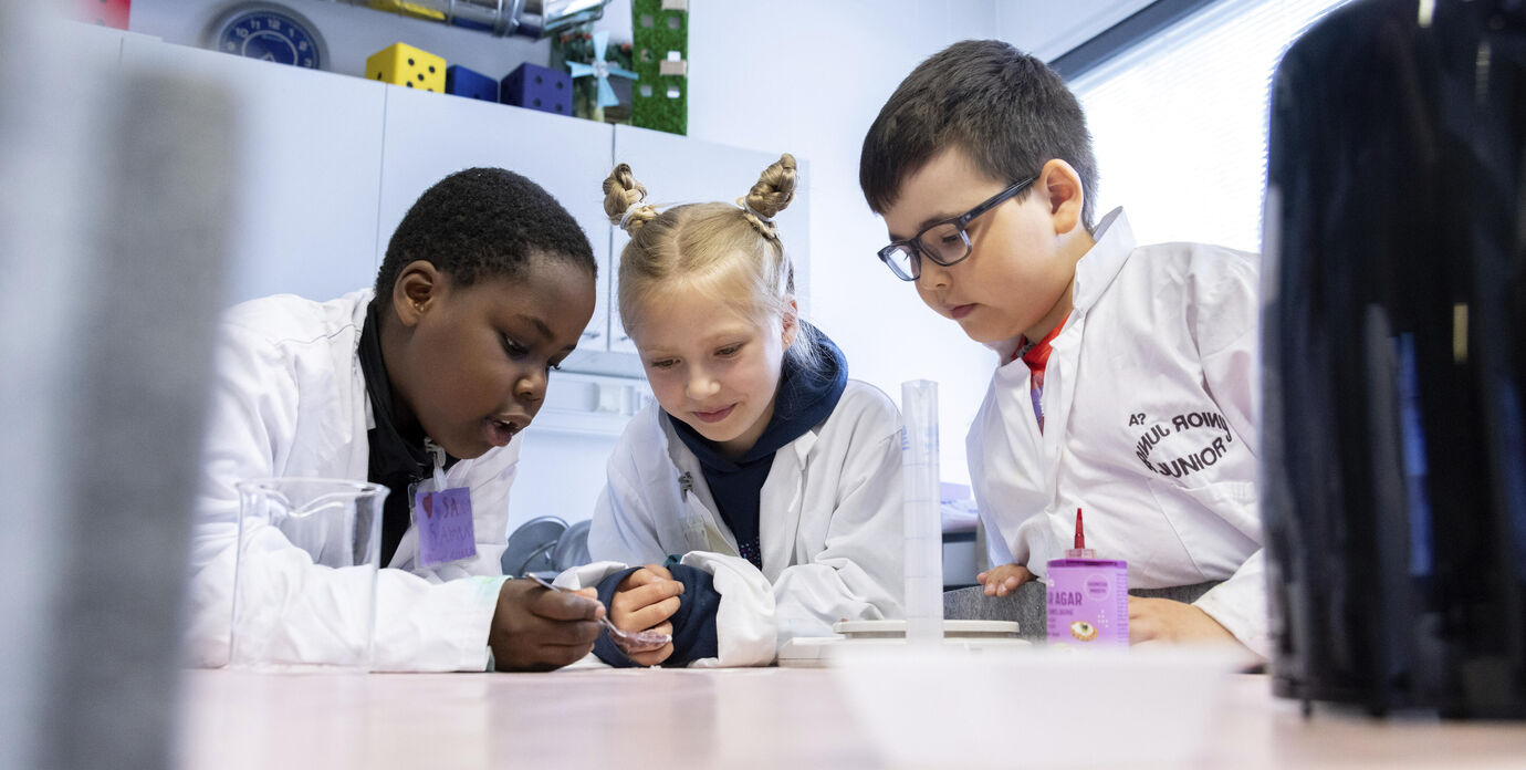 Kolme lasta valkoisissa laboratorio takeissa tekee kokeen luokkahuoneen pöydällä