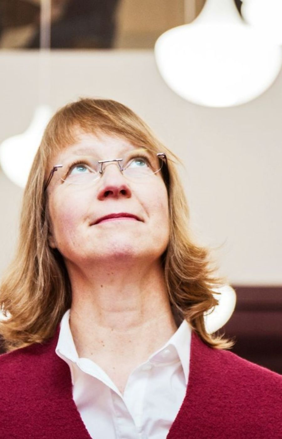 A woman in red cardigan looking up, with white lamps in the background.