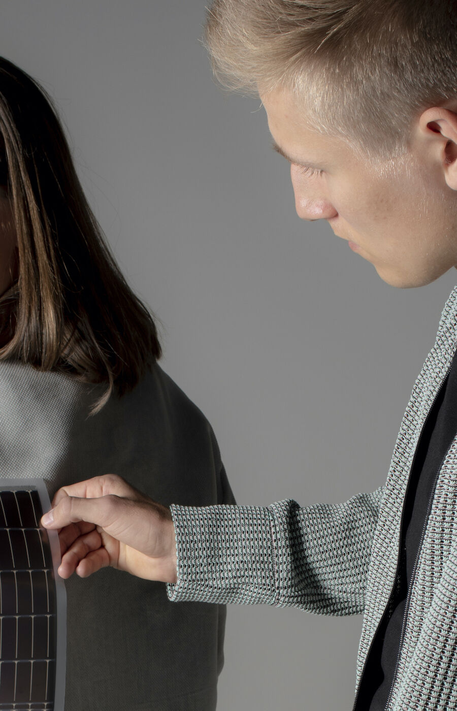 A person attaches a solar panel to the back of another person's light grey jacket.