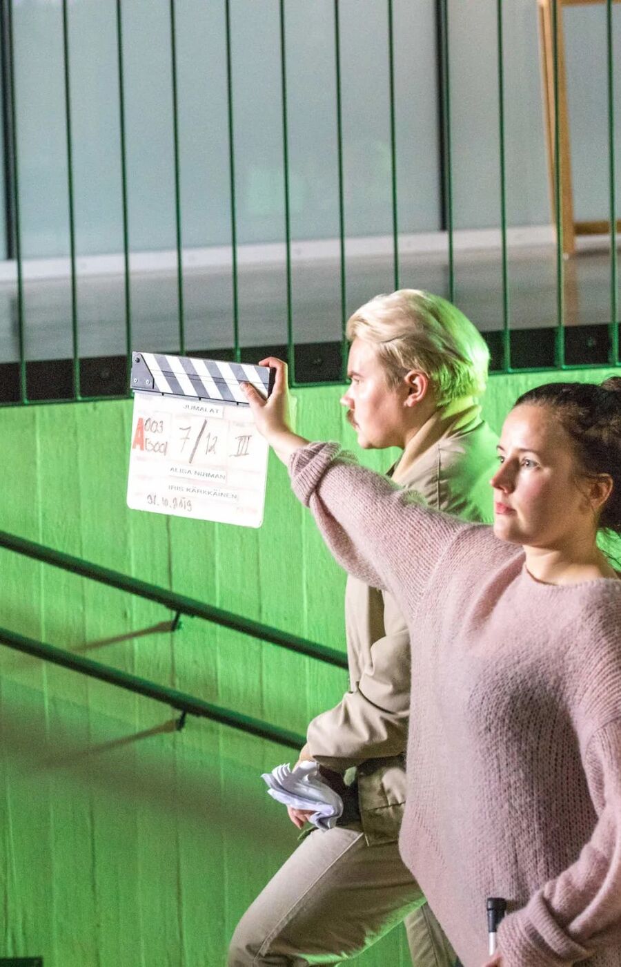 Two people on a film set, one holding a clapperboard. A camera rig is visible on the left. Green wall in the background.