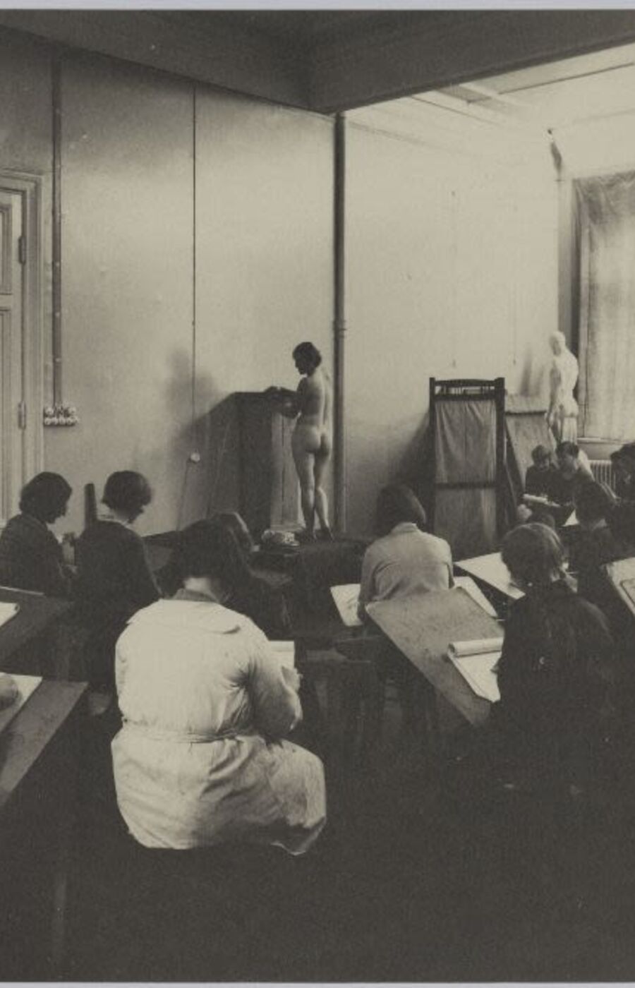 A classroom with students drawing a nude model. The room has large windows and a door on the left.