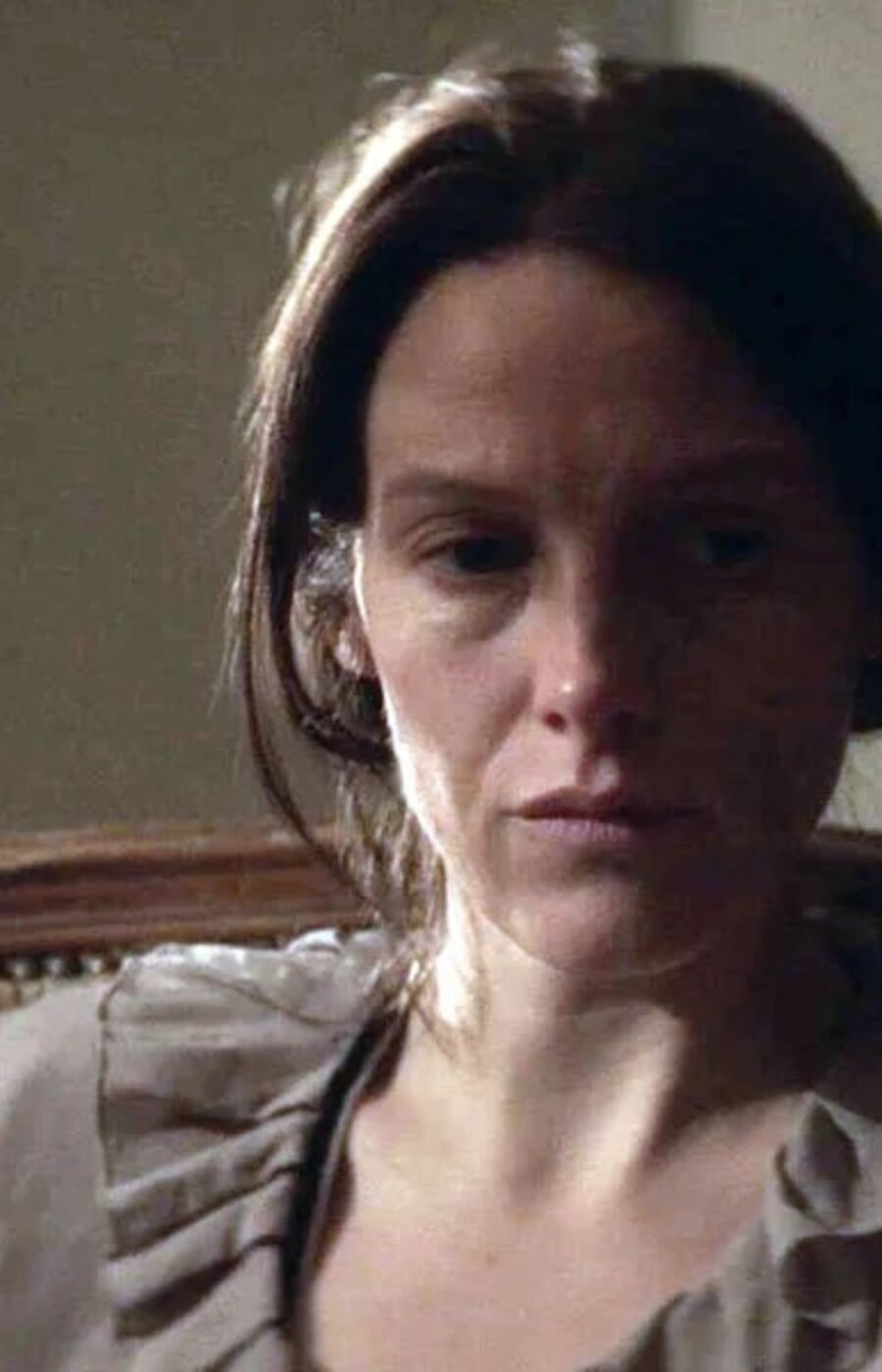 A person with dark hair sits on a vintage chair with a floral pattern. The background is a dimly lit room.