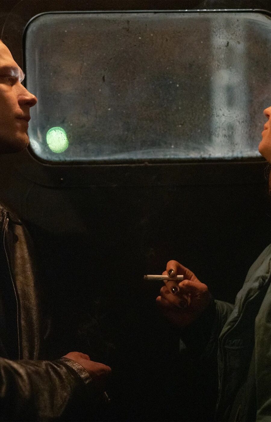Two people standing in front of a window, one wearing a leather jacket and the other a green coat, holding a cigarette.