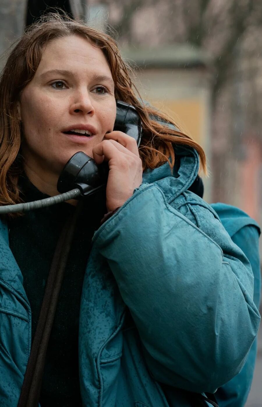 Person in a teal jacket using a public phone booth on a rainy day, with a vintage vehicle in the background.