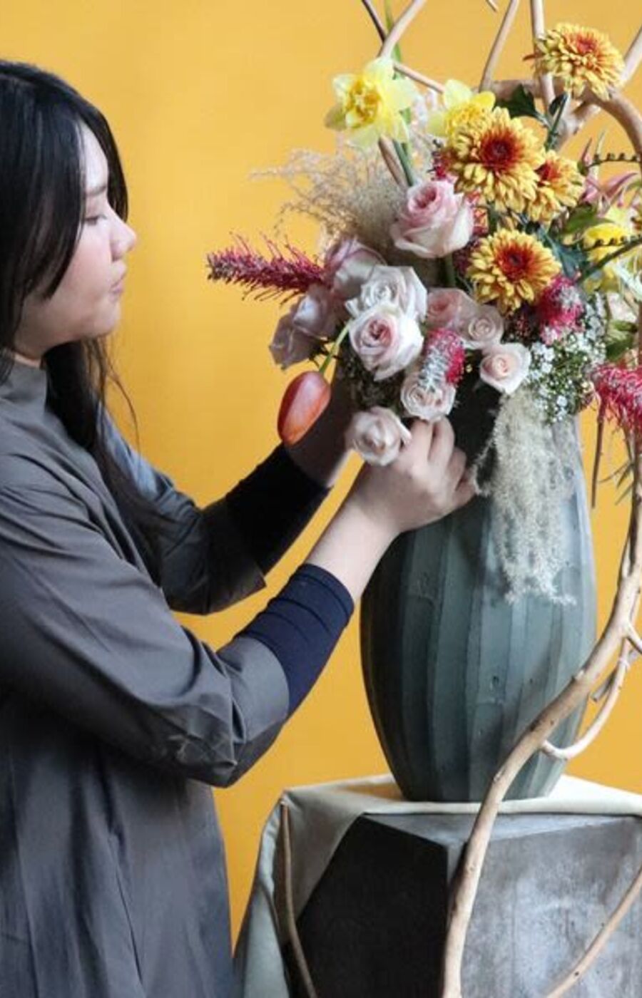 A person arranges a colourful flower bouquet in a green vase on a table with a yellow background.