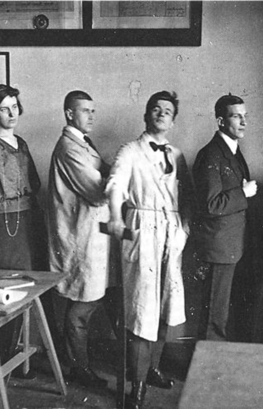 A vintage black and white photo of six people standing in a room with desks and hanging lamps.