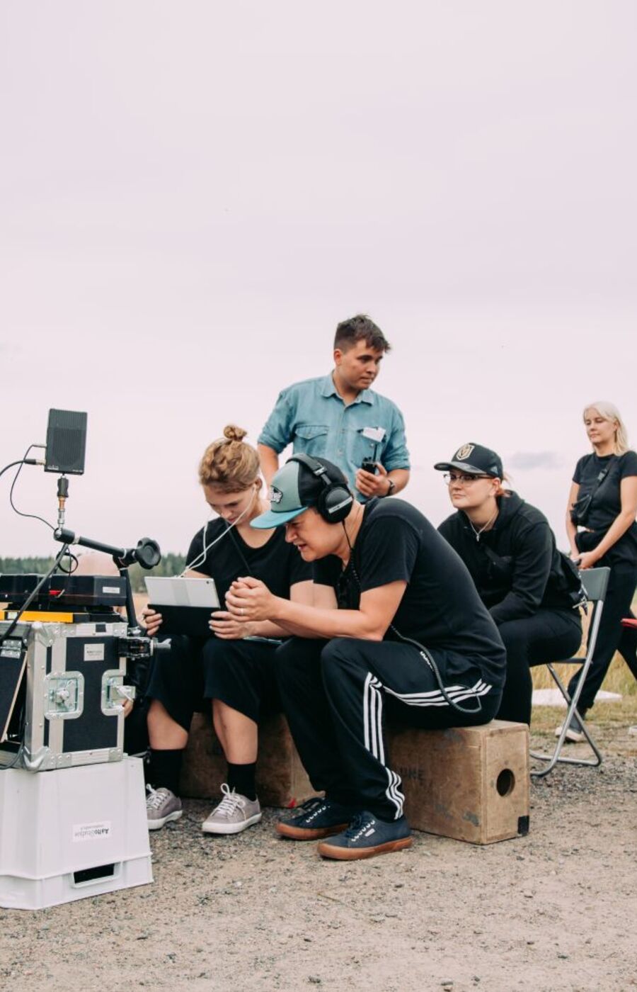 A film crew on location with equipment, including a boom microphone, cameras, and monitors. A white van is in the background.