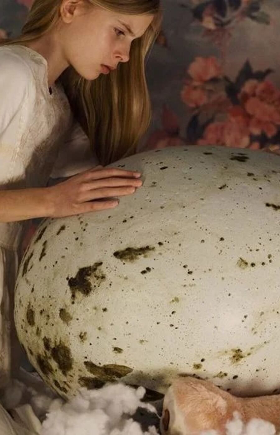 A person in a white dress kneels beside a large speckled egg on a bed with a torn teddy bear and a lamp in the background.