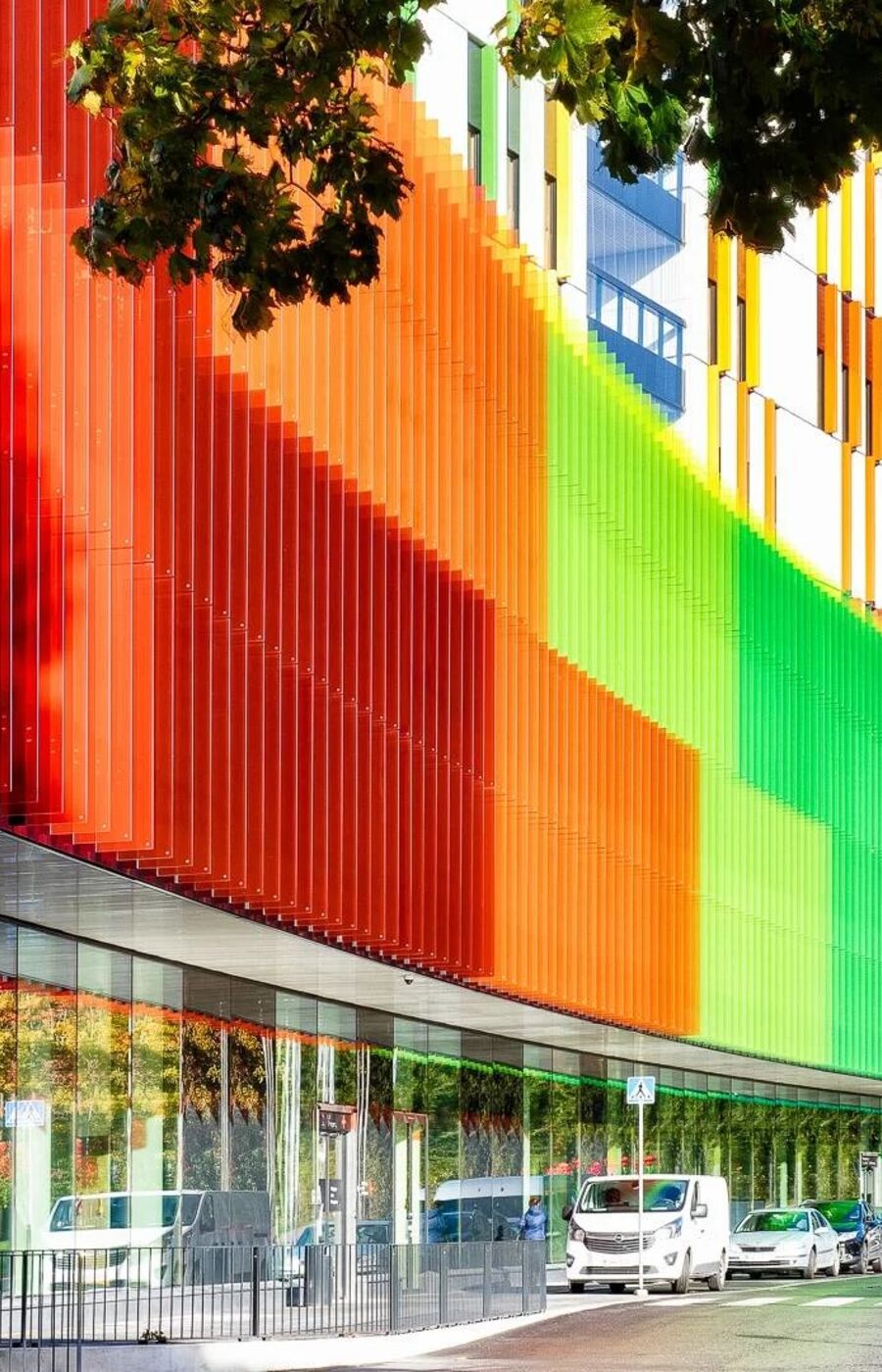 A modern building with a colourful facade featuring vertical panels in red, orange, green, and blue. Cars are parked below.