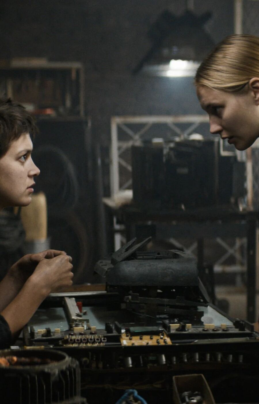 Two people in a workshop, examining a piece of equipment on a table. The room is filled with various tools and machinery.