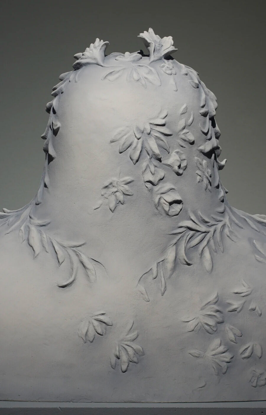 Harmaa veistos, jossa rintakuva on peitetty kukkakuvioilla, lehdet ja kukat nousevat pinnasta.