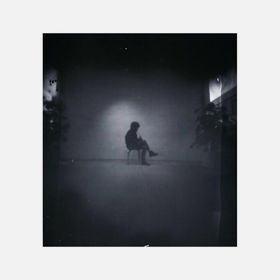 Neulanreikäkameralla kuvattu mustavalkoinen kuva kauempana tuolilla istuvasta henkilöstä. Kuvan reunoilla viitteitä jonkinlaisista kasveista.