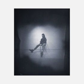 Neulanreikäkameralla otettu mustavalkoinen kuva henkilöstä, joka istuu jakkaralla jalat haarallaan sivuille pitäen kiinni tuolin reunasta, keskeltä.