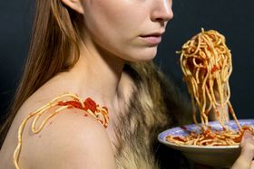 Tiukasti rajattu kuva (näkyvissä kasvot vain nenästä alaspäin hartioihin) pitkähiuksisesta henkilöstä hieman sivusuunnasta, jolla on edessään spagettilautanen. Käsi täynnä spagettia on asetettu suun eteen, mutta suu ei ole auki. Spagettia ja kastiketta on myös  paljaalla olkapäällä. Toinen, taaempi hartia on jonkinlaisen karva-asun peitossa.