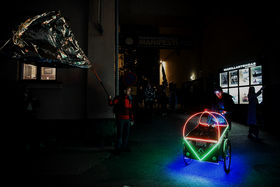 A photo of a perfomance where a person is pushing a two-wheeled cart decorated with neon lights in the street