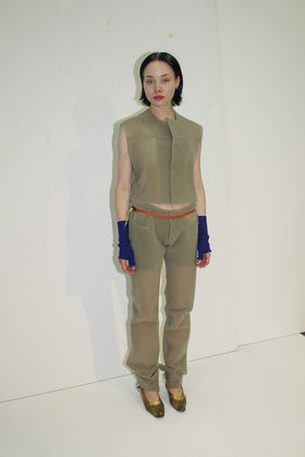 Nainen seisoo vaalean taustan edessä läpikuultavissa vaatteissa ja sinisissä rannekkeissa