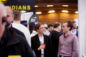 Two people are walking in a crowd and talking to each other. They are looking at each other, and one of them is holding a cup. The other one has a green sticker in his shirt.