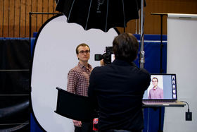 A picture of two men. One of them is standing in front of oval white wall and the other has his back facing the photographer and he is standing behind a camera