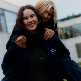 Two people outside, one with dark hair wearing an Aalto University jacket, the other with blonde hair hugging from behind.