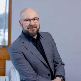 Jaan Praks, jolla on harmaa puvun takki ja musta paita, istuu puisella ikkunanpuitteilla varustetun ikkunan edessä.