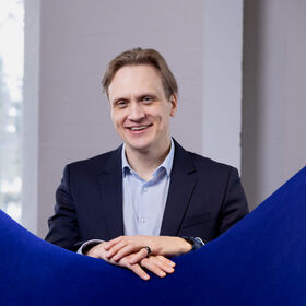 Henkilö tummassa puvussa ja vaaleansinisessä paidassa nojaa kätensä siniseen huonekaluun.