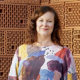 Henkilö, jolla on värikäs abstraktikuvioinen paita, seisoo teksturoidun tiiliseinän edessä.