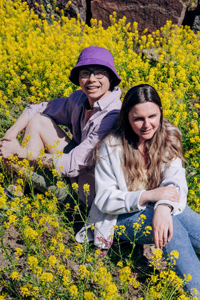 Two Aalto University students sitting outdoors in a field of yellow flowers, one wearing a purple hat and the other in a white jacket.