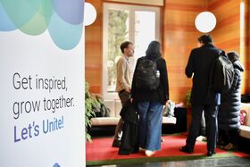 People with backpacks converse in a lobby near a sign that reads, 'Get inspired, grow together. Let's Unite!'.