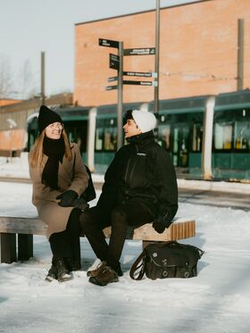 Two people sit on a bench in a snowy urban area with a tram and brick building in the background.
