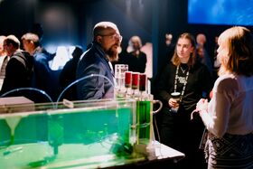 People are gathered around an exhibit that includes green liquid and transparent tubing in a dimly lit room.