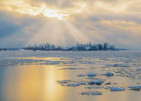 A scenic view of a snowy town with sunlight breaking through cloudy sky, reflecting on icy waters.