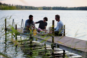 Three people sitting on a wooden jetty by a calm lake, having a picnic with a lantern and drinks.