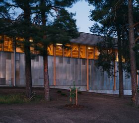 Building exterior with large windows showing wooden beams inside, surrounded by tall pine trees.