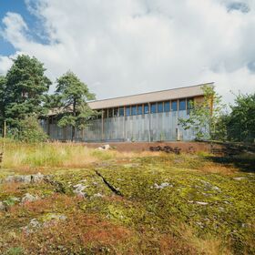 Modern grey storage building with tall windows, surrounded by trees, grass and moss-covered rocky terrain under a partly cloudy sky.