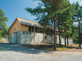 A wooden storage building with windows surrounded by trees. It has a sloped roof and a gravel driveway.