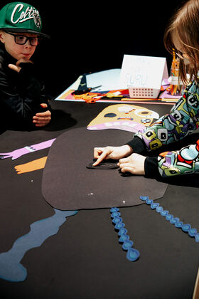 Children crafting colourful paper face art. One child is wearing a patterned jacket and another has a green cap.