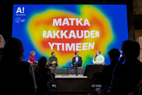 Four people seated on stage with a colourful background reading 'Matka rakkauden ytimeen'. Audience in foreground.