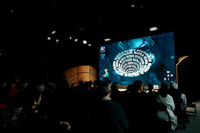 An audience watches a speaker on stage with a large screen displaying graphics and text. The event appears to be educational.
