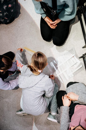 Two children and two adults sitting on the floor, working together with plastic straws and transparent sheets.