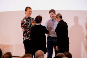 Two men and two women at an event. One man holds a bouquet. Audience visible at the bottom. Background is plain white.