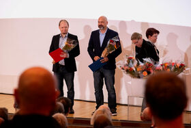 Two men holding flowers and folders stand on stage. Flower bouquets are placed beside them. Audience is visible.
