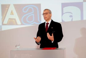 A man in a black suit and red tie presents in front of a projection screen showing large letters 'A' and 'a'.