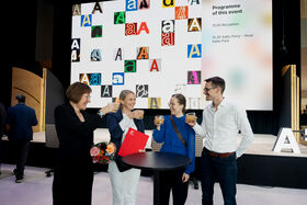 People holding drinks around a round table in front of a large screen with colourful letters. Event programme is displayed.