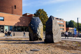Två stora stenar står framför en tegelbyggnad med personer som går förbi. Cyklar är parkerade i närheten.