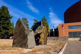 En person i svart rör vid en stor stenskulptur utanför en tegelbyggnad under blå himmel.