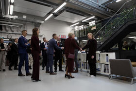 A group of people in business attire are indoors, shaking hands. Industrial setting with staircases and plants.