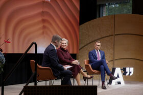 Three people sitting on orange chairs on a stage, with a large screen displaying abstract patterns behind them.