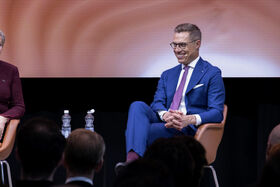 Man in blue suit and pink tie sits on a chair beside a woman in a burgundy outfit. Two water bottles are on a table.
