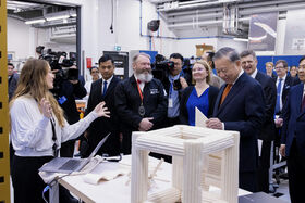 En kvinna i vit skjorta gestikulerar nära en laptop. Flera personer i formella kläder observerar henne i en labbmiljö.